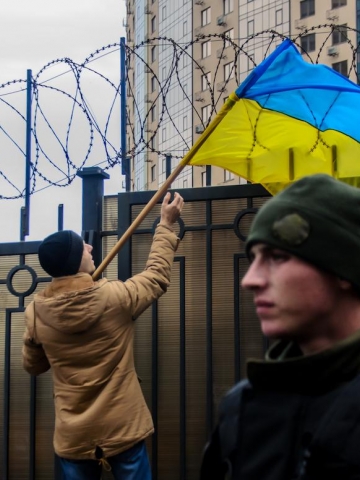 A man holds a Ukrainian flag near a barbed wire divider. On the other side of the divider, a Russian flag is visible.